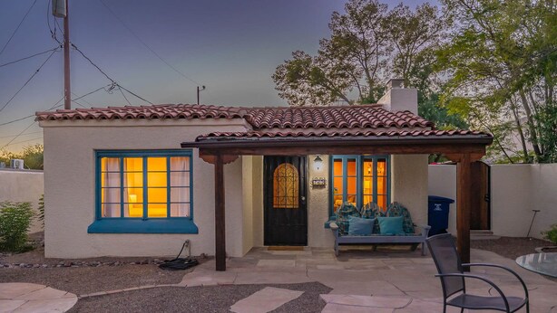 A small house with a tiled roof, a porch with a bench, and a patio area with a chair.