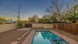 A rectangular swimming pool with a blue tiled edge, surrounded by a concrete deck with stepping stones leading to a gated entrance.