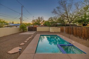 A rectangular swimming pool with a blue tiled edge, surrounded by a concrete deck with stepping stones leading to a gated entrance.