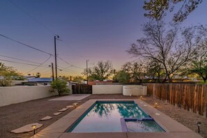 A rectangular swimming pool with a tiled deck, surrounded by a wooden fence, and a clear sky above.