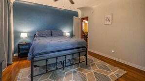 A bedroom with a bed, two bedside tables with lamps, a ceiling fan, a patterned rug, and a door leading to another room.