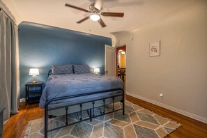 A bedroom with a bed, two bedside tables with lamps, a ceiling fan, a patterned rug, and a door leading to another room.