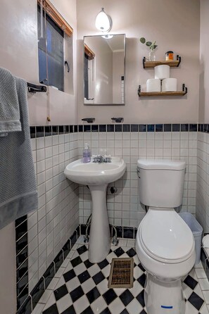A bathroom with a sink, toilet, and mirror. There is a towel rack and a shelf with toiletries.