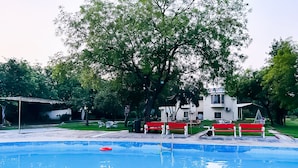 A swimming pool with a red and white bench area, surrounded by trees and a building. - Roomshala 131 Willow Woods Farmhouse (Sohna)