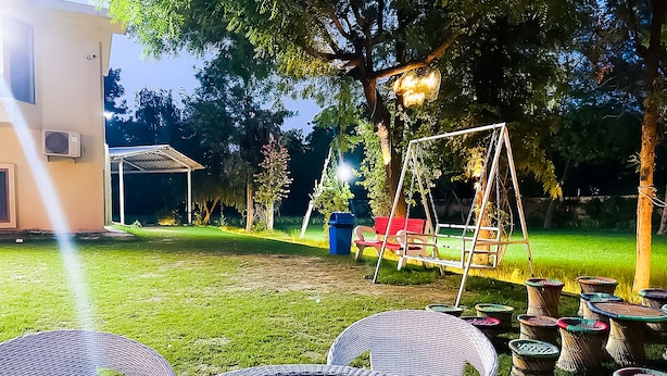 A backyard with a glass table, wicker chairs, a swing set, and a small shed. - Roomshala 131 Willow Woods Farmhouse (Sohna)