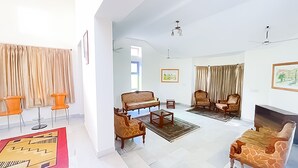A spacious living room with a sofa set, armchairs, and a coffee table. There are curtains on the windows, and a painting is visible on the wall. - Roomshala 131 Willow Woods Farmhouse (Sohna)
