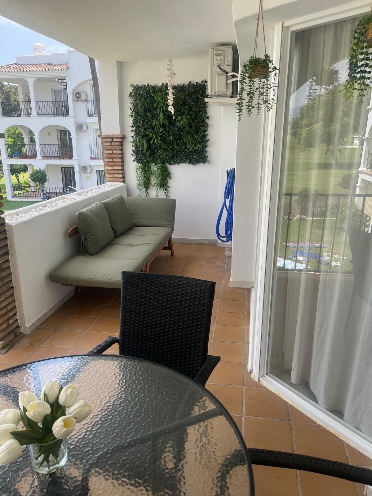 Ein Balkon mit einer Couch, einem Tisch mit Blumenvase und einer hängenden Pflanze.