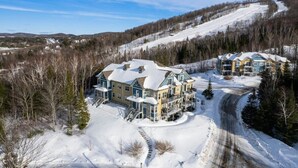 Un edificio grande de varios pisos con techo cubierto de nieve, rodeado de árboles y una pista de esquí al fondo.