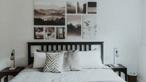 Un dormitorio con una cama, dos mesitas de noche, una obra de arte en la pared y una mesita con una lámpara.