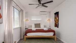 Un dormitorio con cama, ventilador de techo, ventana con persianas y aire acondicionado de pared.