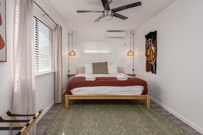 Une chambre à coucher comprenant un lit, un ventilateur de plafond, une fenêtre avec des stores et un climatiseur fixé au mur.