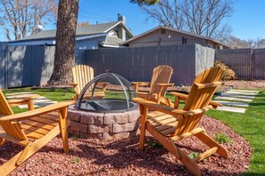 Un patio trasero con un hogar de piedra rodeado de sillas Adirondack de madera, una terraza de madera y un jardín cercado.