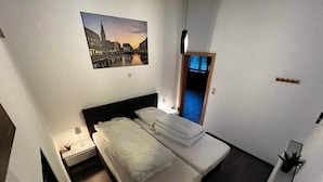 A modern bedroom with a bed, a nightstand, a lamp, a painting of a cityscape, and a small plant. - Ecolodge Loft, 180qm Raumwunder für bis zu 8 Personen, 5km zu Nordsee / Büsum (Oesterdeichstrich)