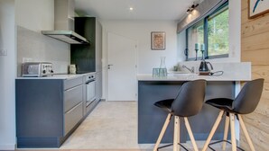 A modern kitchen with a built-in stove, a toaster, and a sink. There are two bar stools and a window with a view of trees. - Alptitude Beauvallon Petit - stylish and spacious property for 6 people (Morzine)