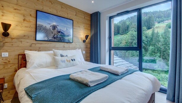 A bedroom with a large bed, wooden walls, a painting, and a view of a forest through a large window. - Alptitude Beauvallon Petit - stylish and spacious property for 6 people (Morzine)