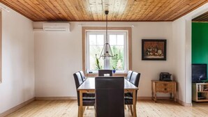 Un coin repas avec une table et des chaises en bois, un luminaire suspendu, une fenêtre donnant sur un espace verdoyant et un téléviseur posé sur un meuble.