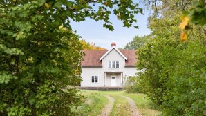 Une maison avec un toit recouvert de tuiles rouges, des murs blancs et une porte d’entrée. Un chemin mène à la maison, qui est entourée de verdure.