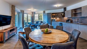 A modern living room with a wooden dining table, comfortable seating, a flat-screen TV, and a large window offering a scenic view.
