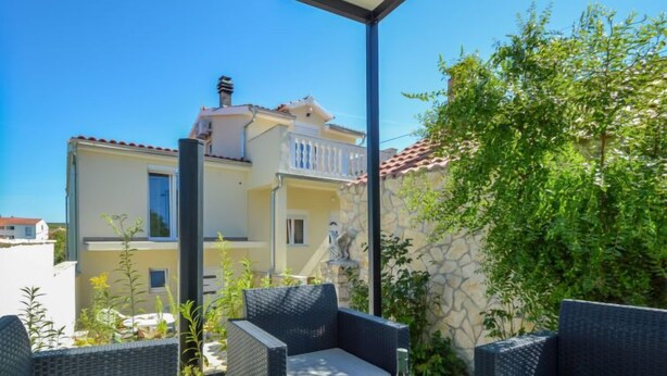 A covered outdoor seating area with wicker furniture, a table with a bottle and glasses, and a view of a building with a balcony. - Vacation home Aurora by Interhome (Skradin)