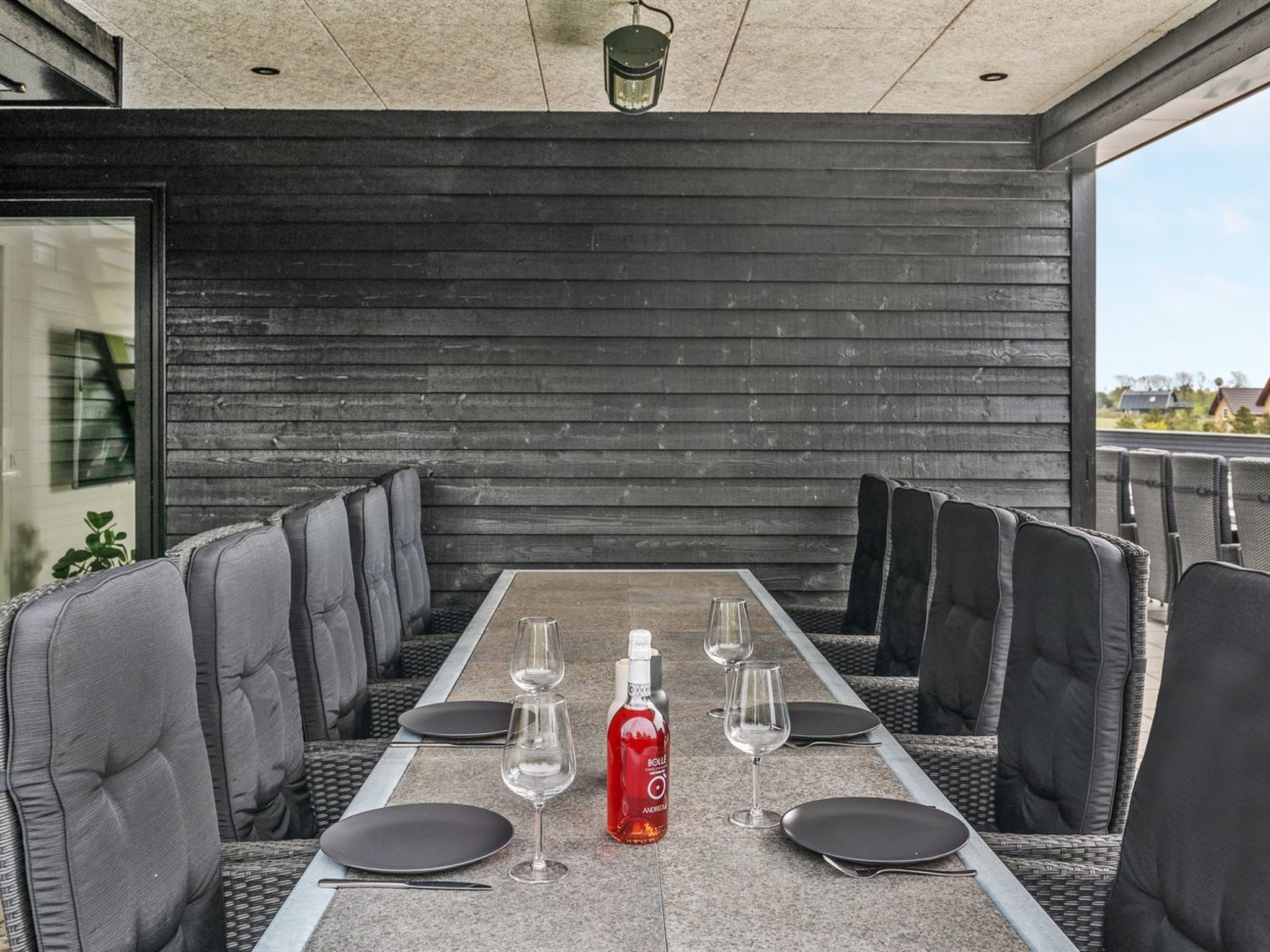 An outdoor dining area with a long table, black cushioned chairs, and a bottle of red wine.