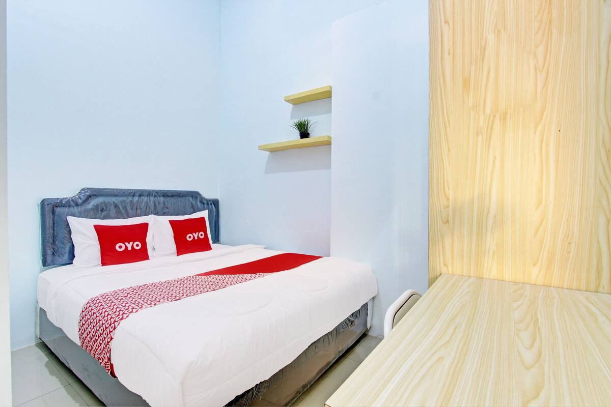 A bedroom with a bed, pillows, a bedside table, a small shelf with a plant, and a wooden panel.