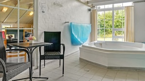 A modern bathroom with a large bathtub, a window with a view of greenery, and a seating area with a table and chairs.