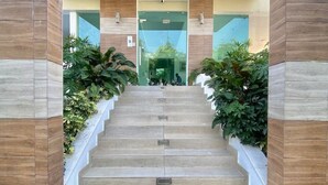 Una escalera conduce a la entrada de un edificio con puerta de vidrio, flanqueada por plantas y paneles de madera.