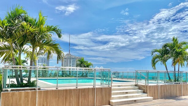 Terraza de madera con barandas de vidrio que da a una piscina, palmeras y un cielo azul despejado.