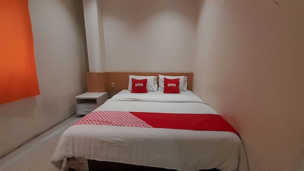 Une chambre d’hôtel avec un lit, deux oreillers ornés du mot « OYO », une couvre-lit blanche et rouge, une table de chevet blanche et une tête de lit en bois.