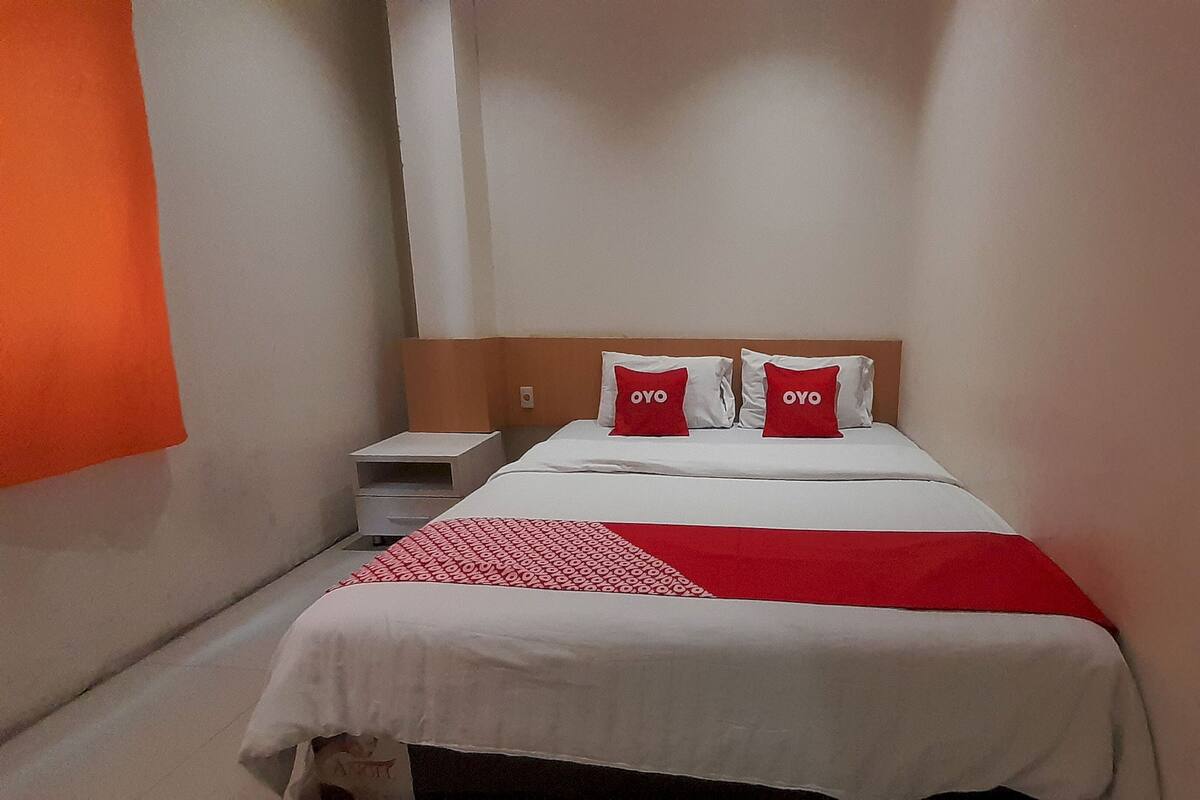 Ein Hotelzimmer mit einem Bett, zwei Kissen mit der Aufschrift "OYO", einer weiß-roten Tagesdecke, einem weißen Nachttisch und einem Holzkopfbrett.