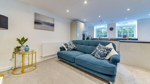 Ein modernes Wohnzimmer mit einem blauen Sofa, einem kleinen runden Tisch mit einer Pflanze und einer Küchenzeile im Hintergrund.