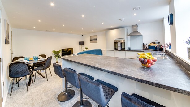 A modern kitchen with a central island, bar stools, and a dining area with a table and chairs. - Stunning Executive 2 Bedroom House (Worksop)