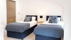 A room with two beds, each with a dark blue headboard and matching bedspread. There is a wooden door on the left and a nightstand with a lamp between the beds. - Stunning Executive 2 Bedroom House (Worksop)