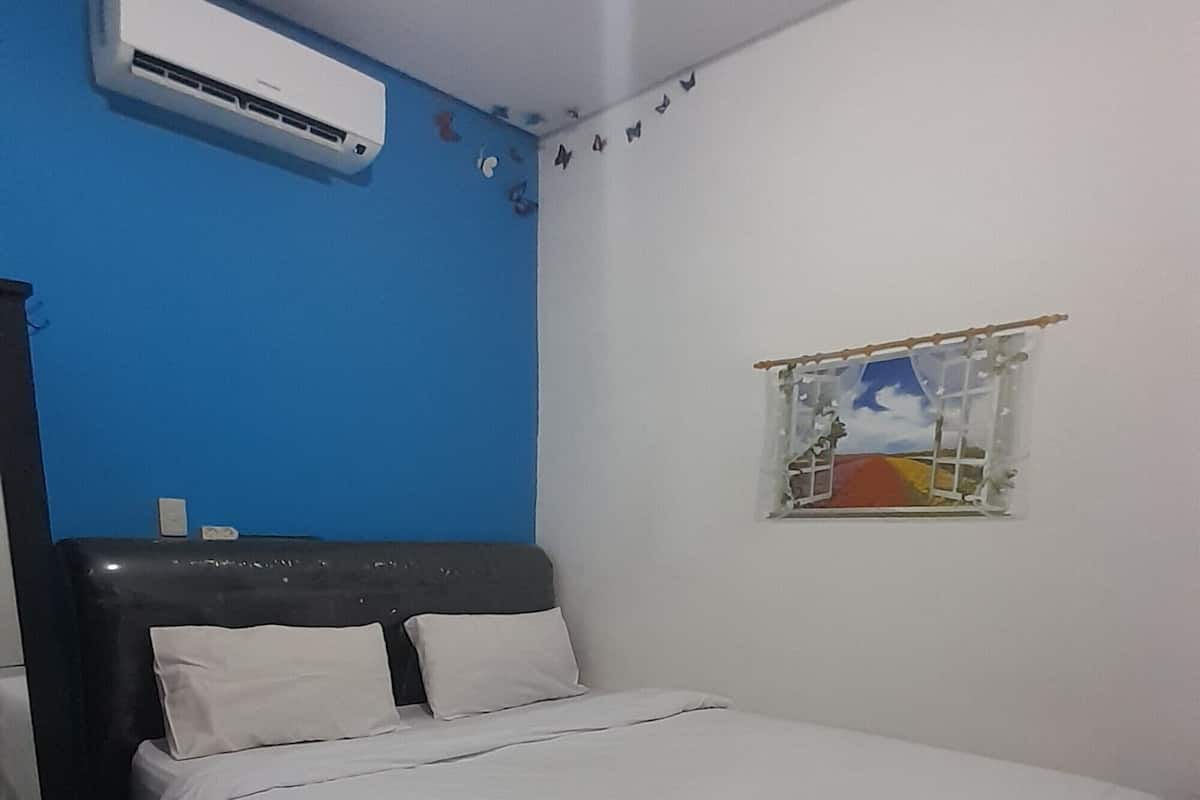 A bedroom with a bed, white bedding, an air conditioning unit, a blue wall, and a small window with a view.