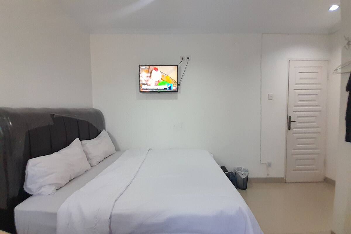 A bedroom with a bed, a TV mounted on the wall, a door, and a trash bin.