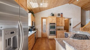 A modern kitchen with stainless steel appliances, wooden cabinets, and a granite countertop. - Hot Tub, Game Room, Large Deck w/ Mountain Views, 4BDR | Paw Print by AvantStay (Big Bear Lake)
