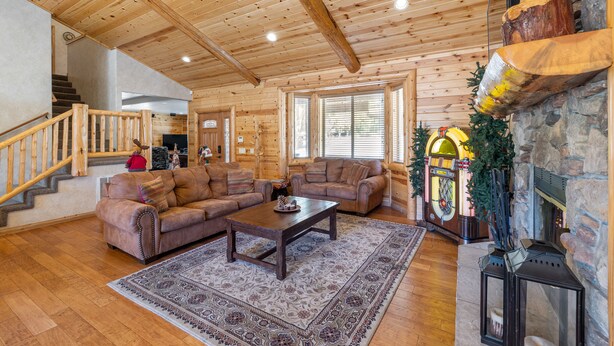A spacious living room with wooden floors, a large sofa set, a coffee table, and a stone fireplace. - Hot Tub, Game Room, Large Deck w/ Mountain Views, 4BDR | Paw Print by AvantStay (Big Bear Lake)
