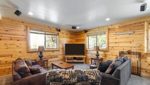 A cozy cabin interior with wooden walls, a flat-screen TV, two sofas, and a patterned ottoman. - Hot Tub, Game Room, Large Deck w/ Mountain Views, 4BDR | Paw Print by AvantStay (Big Bear Lake)