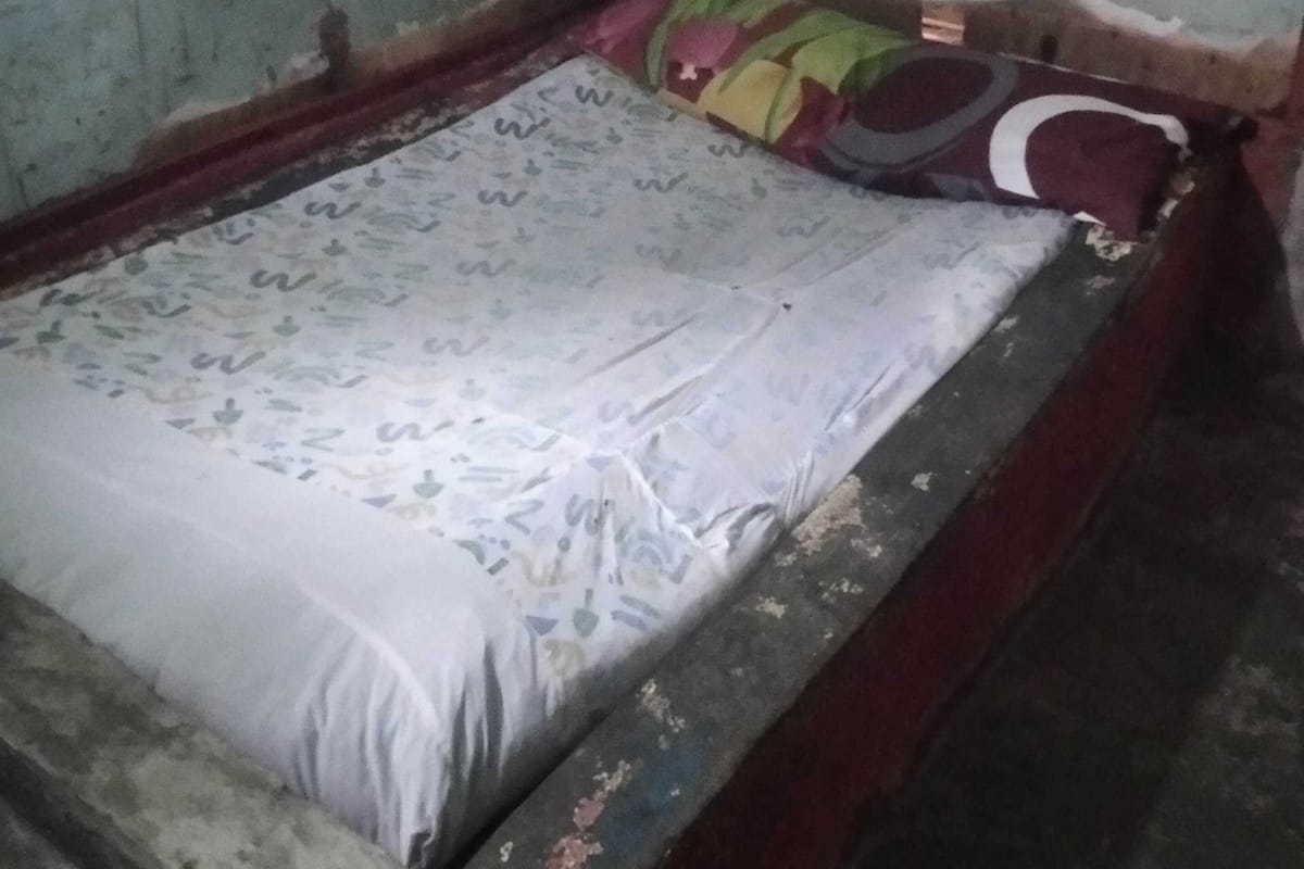 Une petite pièce faiblement éclairée, avec un lit simple, un matelas usé et un cadre en bois sobre.