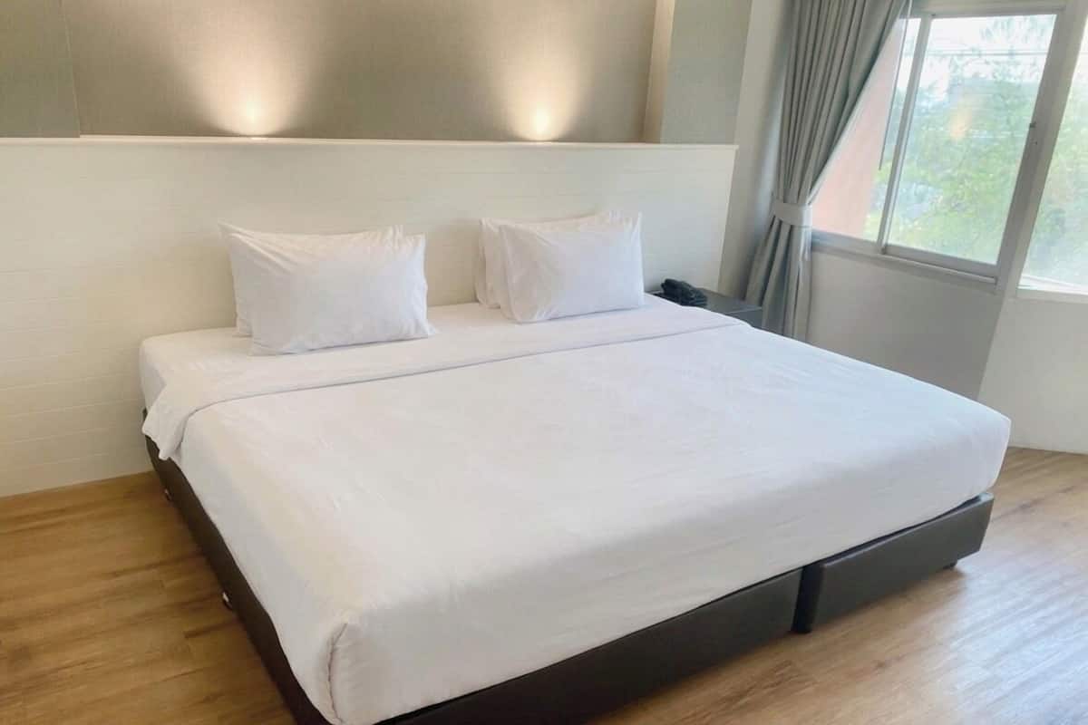 Una cama grande con sábanas blancas, piso de madera y una ventana con cortinas.