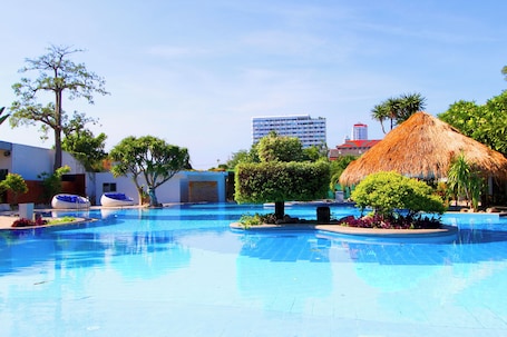 A swimming pool with a thatched-roof structure, surrounded by greenery and modern buildings in the background.. Coco Beach Hotel Jomtien Pattaya