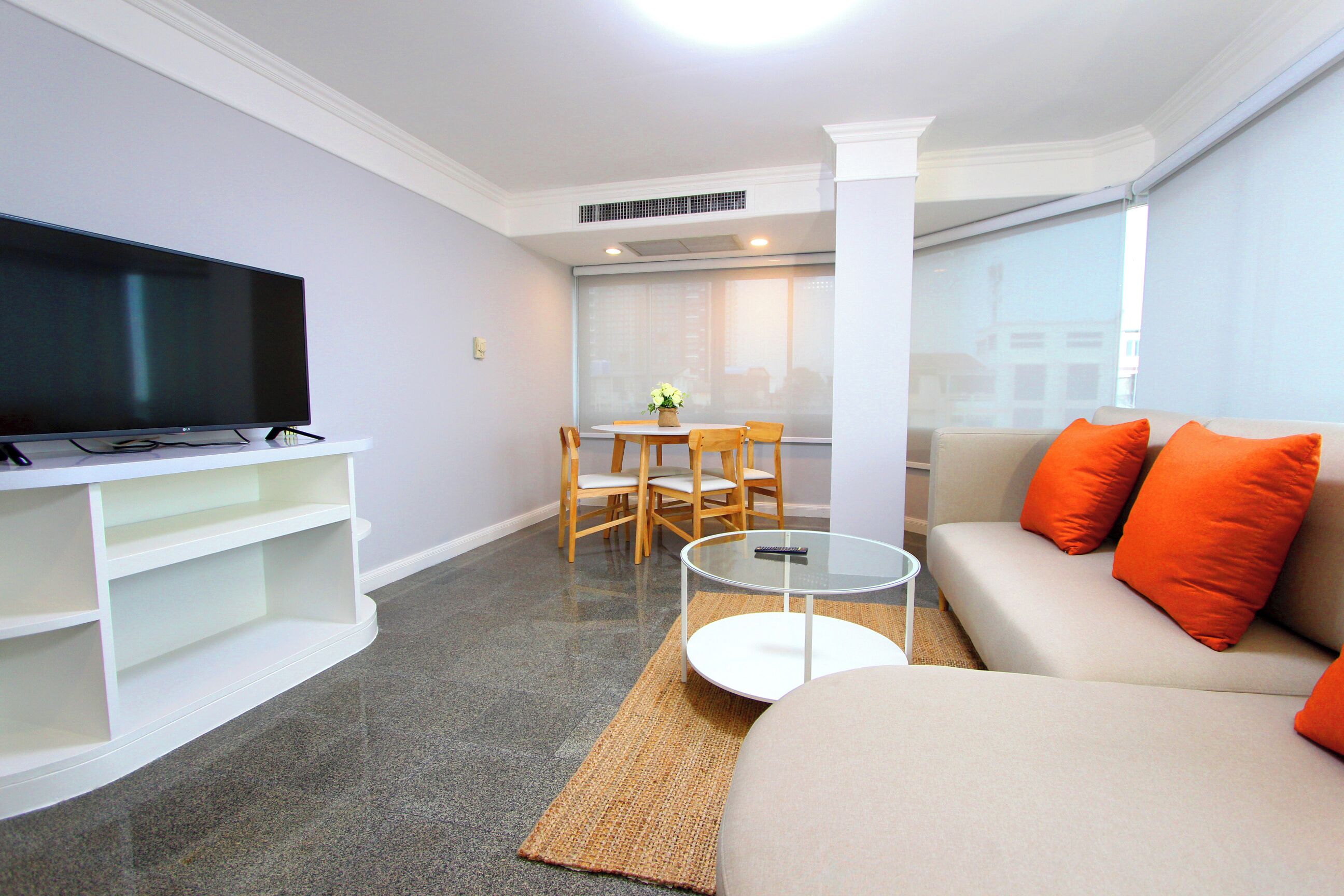A modern living room with a flat-screen TV, a sofa with orange cushions, a glass coffee table, and a dining area with a table and chairs.