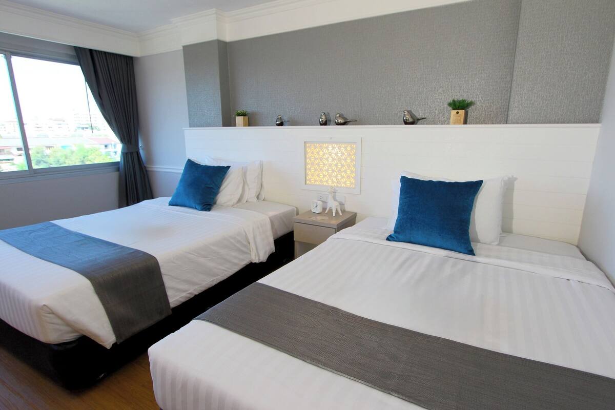 Habitación de hotel con dos camas, una mesita de noche, un estante en la pared con objetos decorativos y una ventana con cortinas.