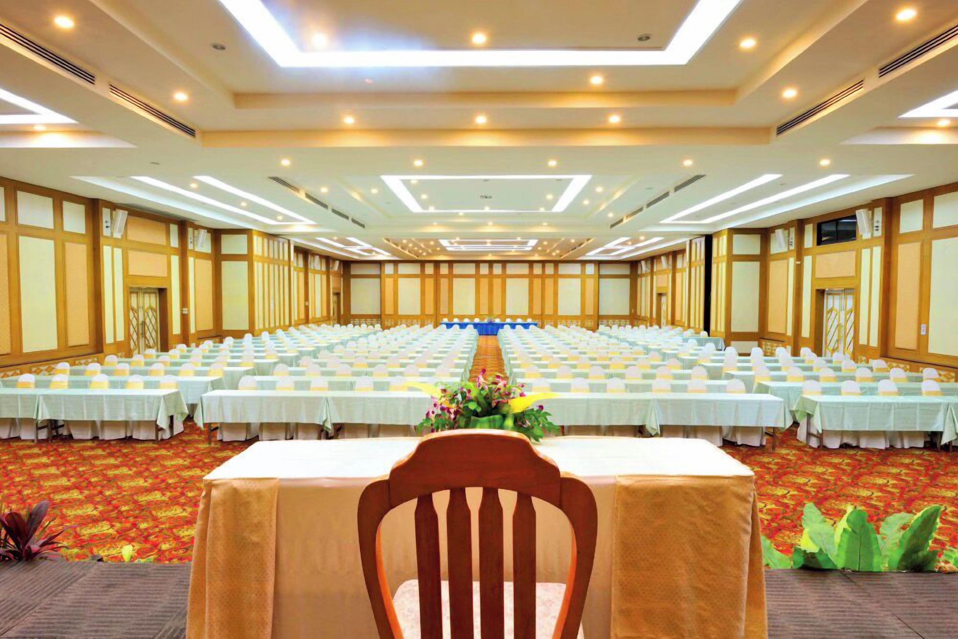 Un amplio salón de conferencias con filas de mesas y sillas, un escenario frontal y suelo alfombrado.