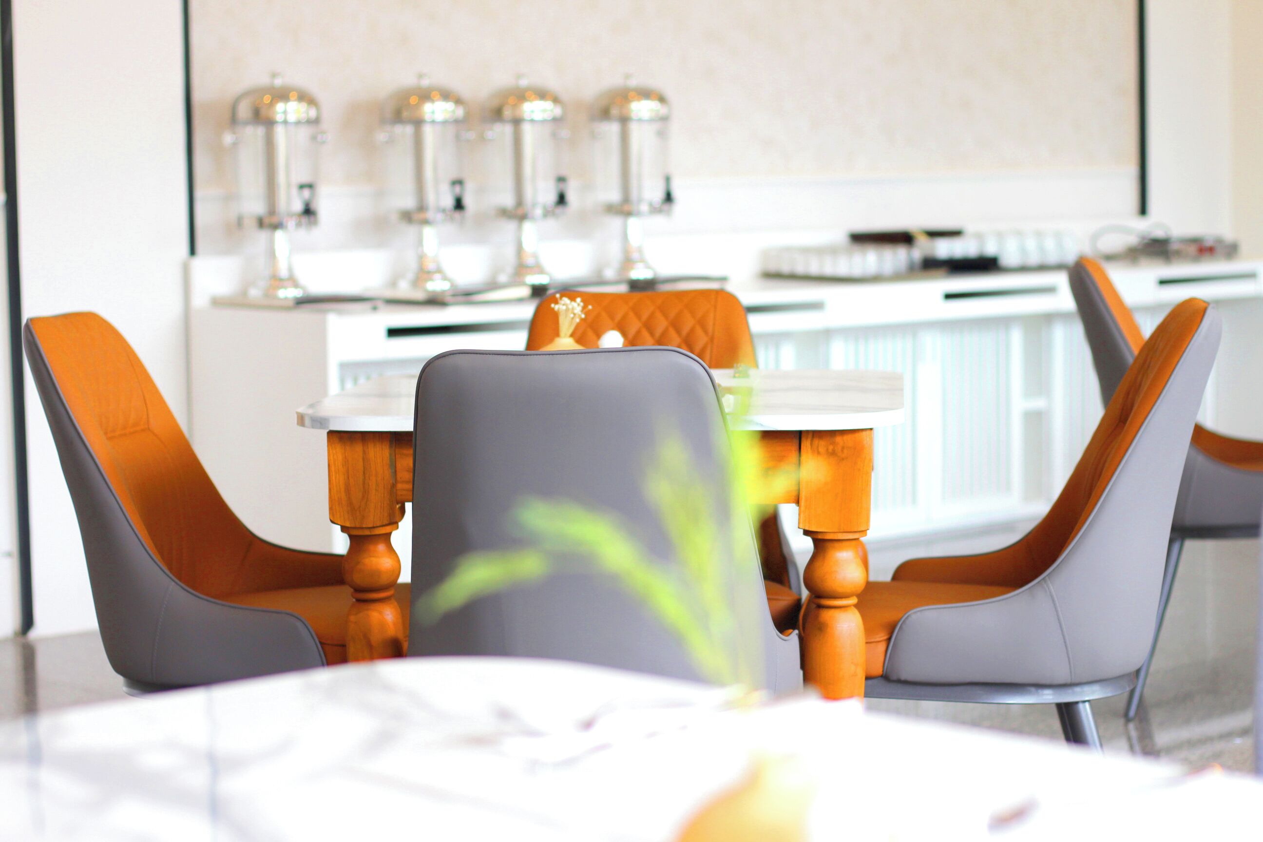 A modern dining area with a white table, grey and orange chairs, and a kitchen in the background with stainless steel appliances.
