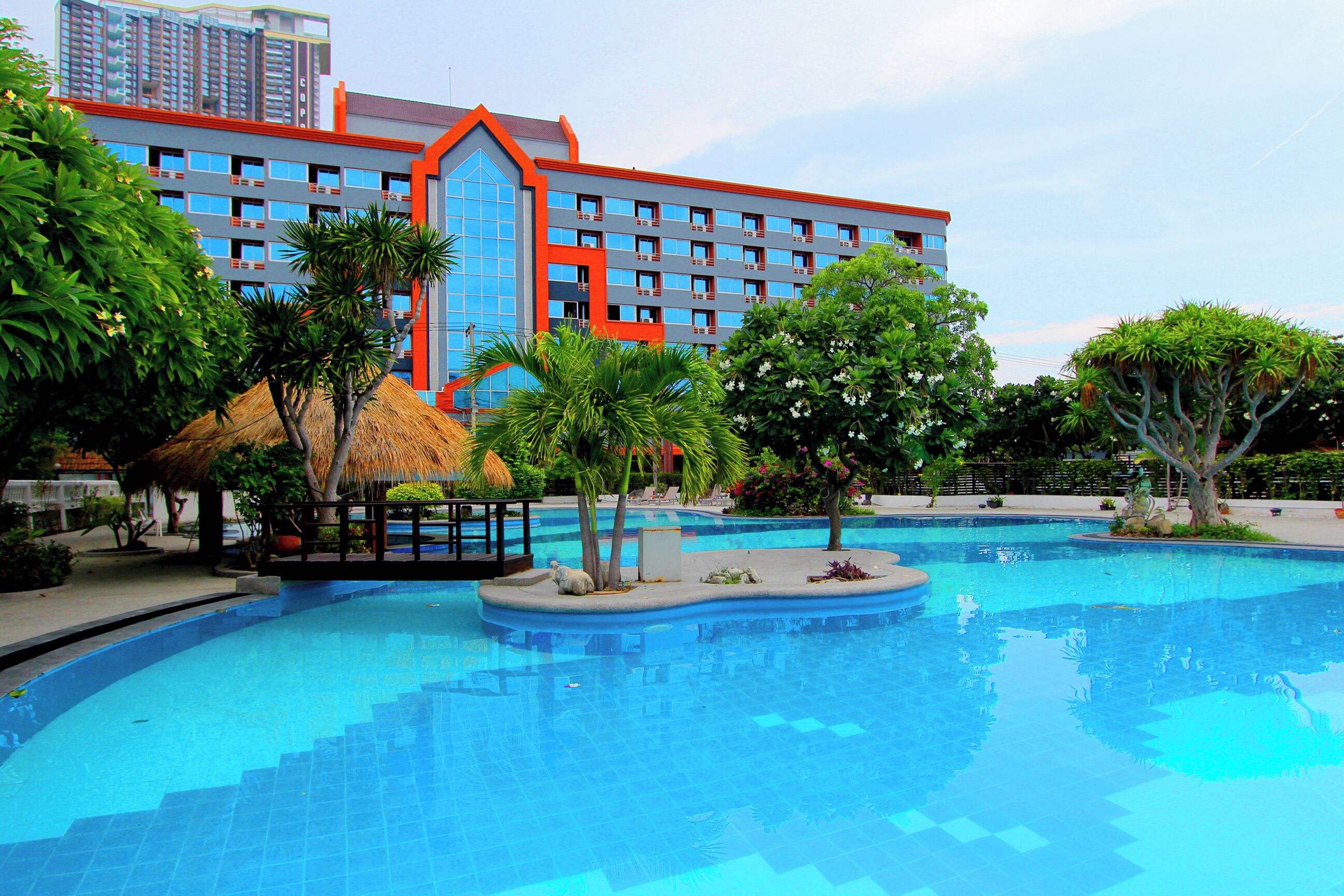 A swimming pool with a thatched-roof structure, surrounded by tropical plants and a hotel building with multiple floors and balconies.