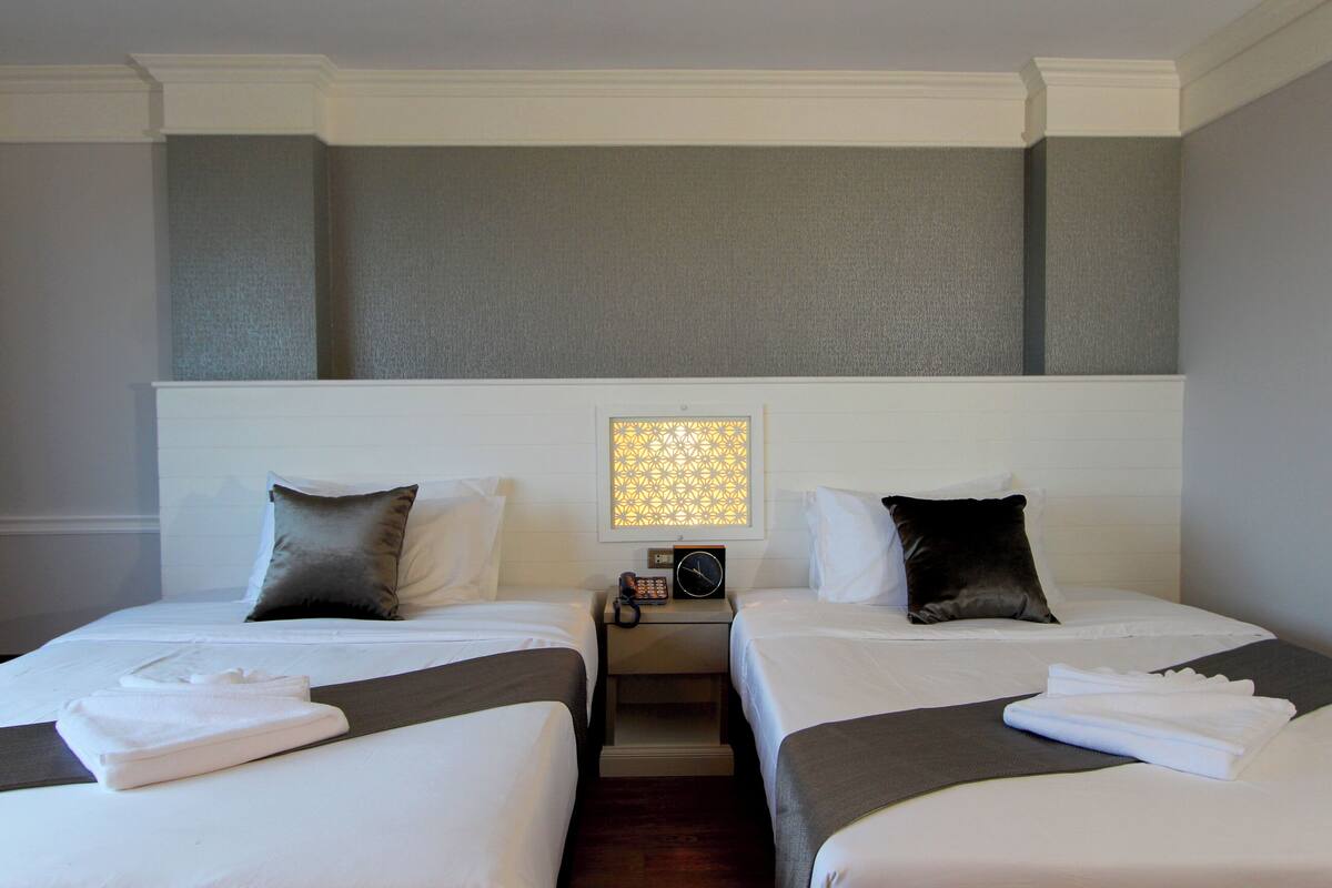 Habitación de hotel con dos camas, una mesita de noche con lámpara y un panel decorativo en la pared.
