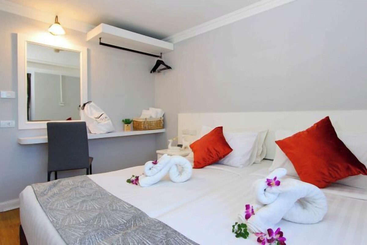 Habitación de hotel con cama, una silla, un espejo, estante con artículos de aseo y toallas dispuestas sobre la cama.