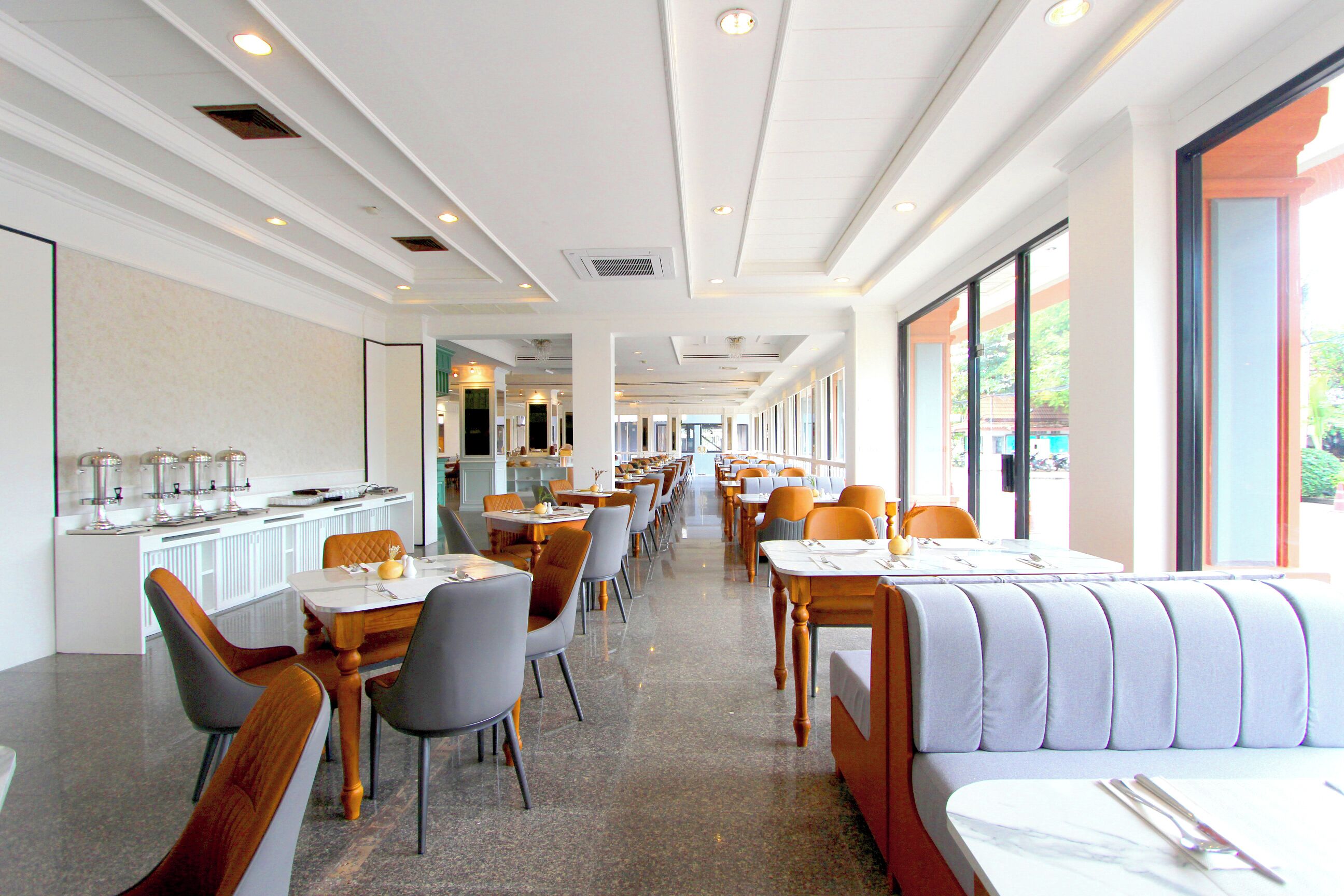 A spacious hotel dining area with multiple tables and chairs, a long counter with a buffet, and large windows providing natural light.
