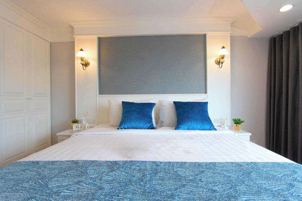 Habitación de hotel con una cama grande, dos lámparas fijadas a la pared, un reloj y una planta pequeña en la mesita de noche.